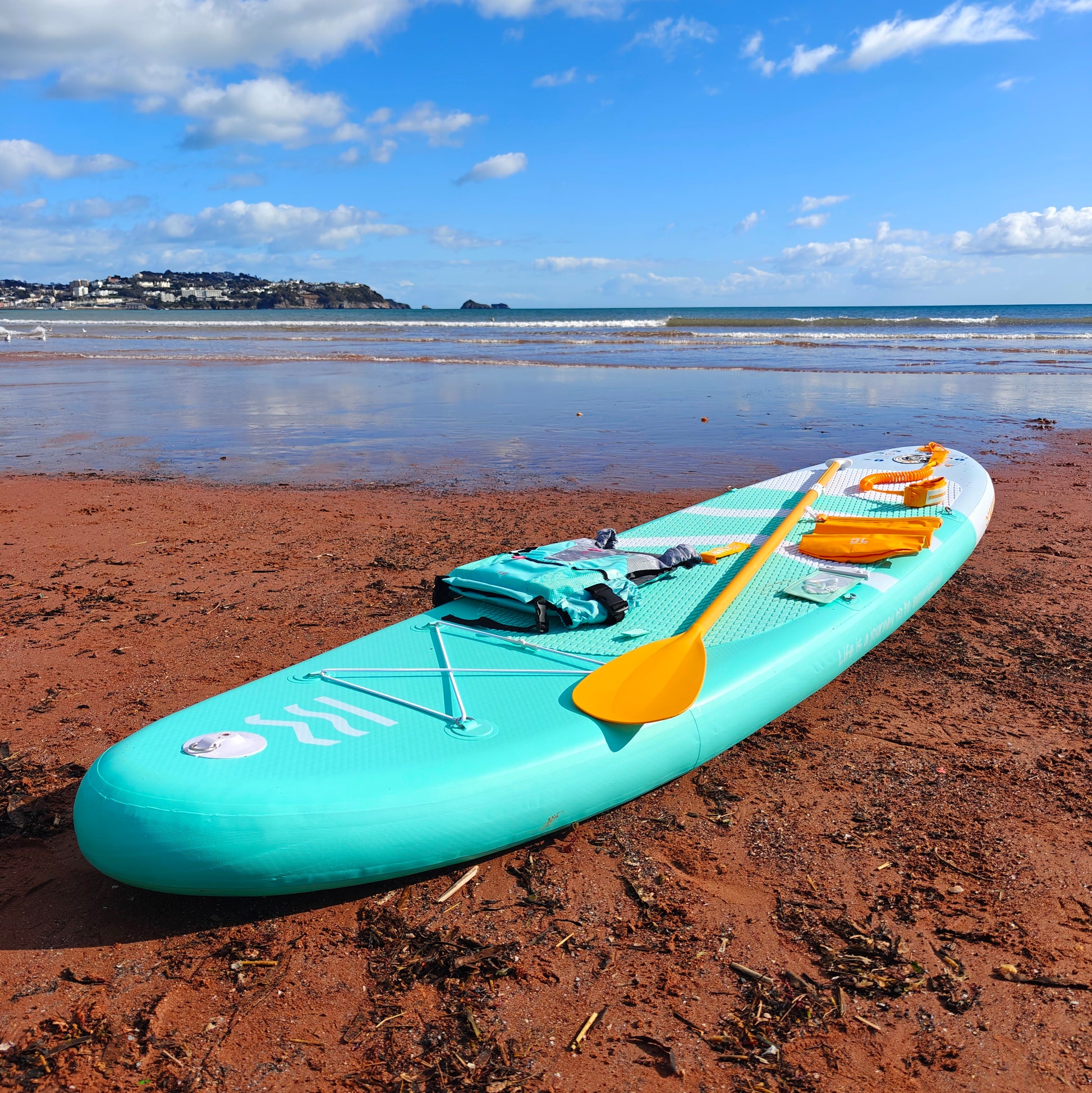 Turquoise paddleboard with a yellow paddle on a sandy beach with ocean and sky in the background